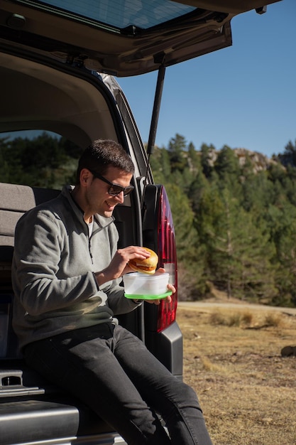 Giovane uomo seduto in un camper che mangia un hamburger vegano e si gode il paesaggio