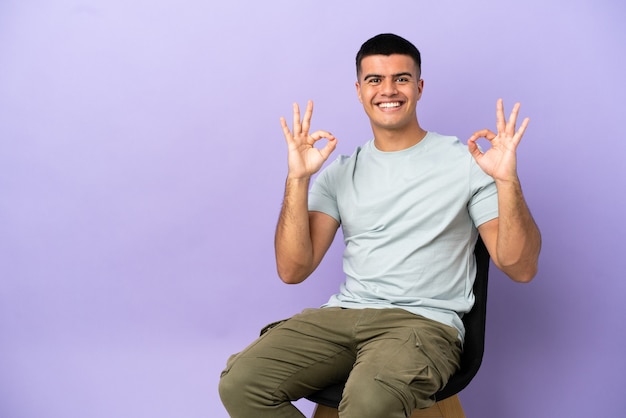 Young man sitting on a chair over isolated background showing an ok sign with fingers