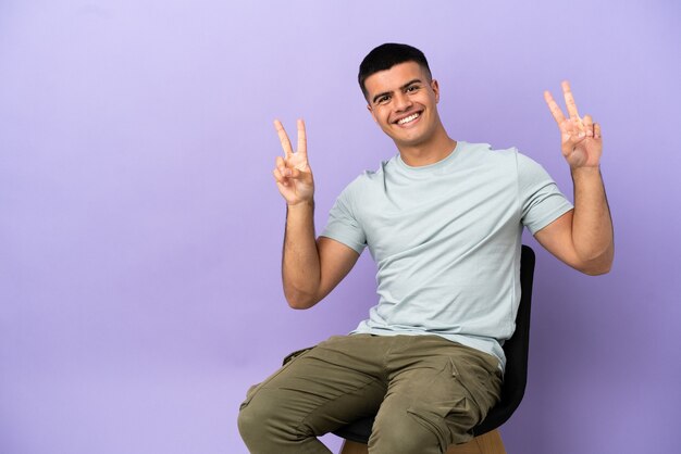 Young man sitting on a chair over isolated background showing victory sign with both hands