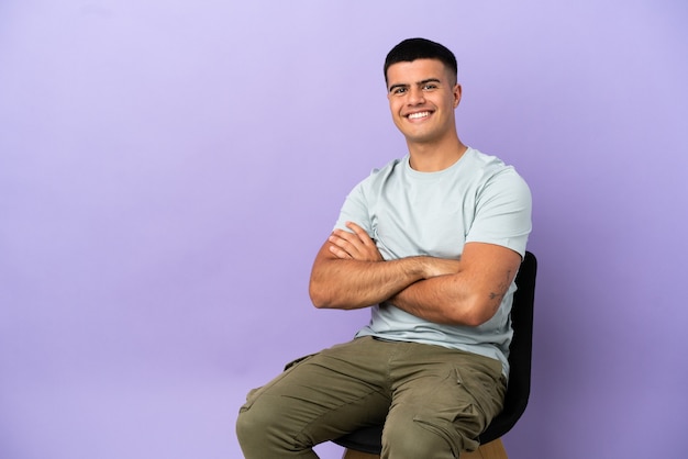 Young man sitting on a chair over isolated background with arms crossed and looking forward