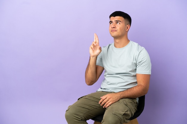 Young man sitting on a chair over isolated background with fingers crossing and wishing the best