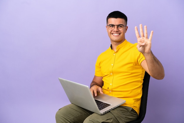 Young man sitting on a chair with laptop happy and counting four with fingers