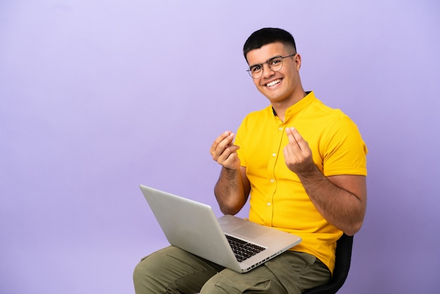 Young man sitting on a chair with laptop making money gesture