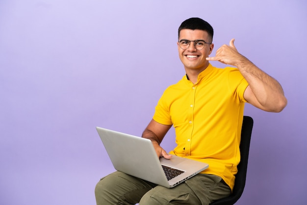 Young man sitting on a chair with laptop making phone gesture. Call me back sign