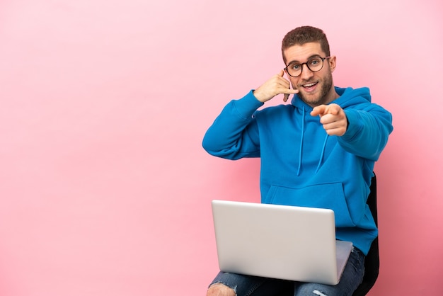 Giovane che si siede su una sedia con il computer portatile che fa il gesto del telefono e che indica la parte anteriore