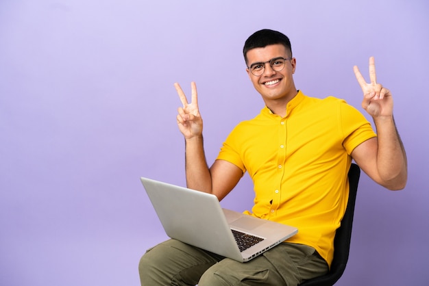 Young man sitting on a chair with laptop showing victory sign with both hands