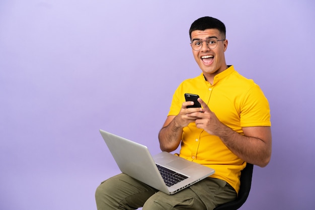 Young man sitting on a chair with laptop surprised and sending a message
