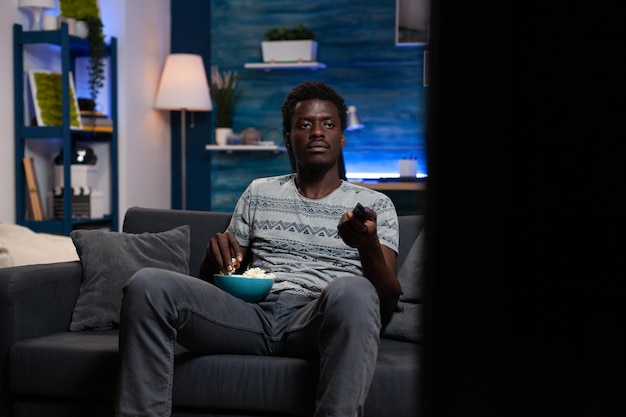 Photo young man sitting on couch looking at television watching entertainment film enjoying lesiure activity in living room after busy day. guy holding popcorn bowl drinking beer during comedy movie