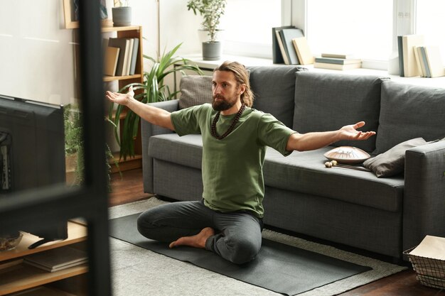 Foto giovane seduto su un tappetino da ginnastica davanti alla tv guarda il programma e fa yoga a casa