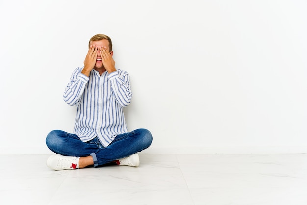 Young man sitting on the floor covers eyes with hands, smiles broadly waiting for a surprise