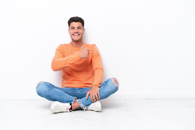 Young man sitting on the floor isolated on white background giving a thumbs up gesture