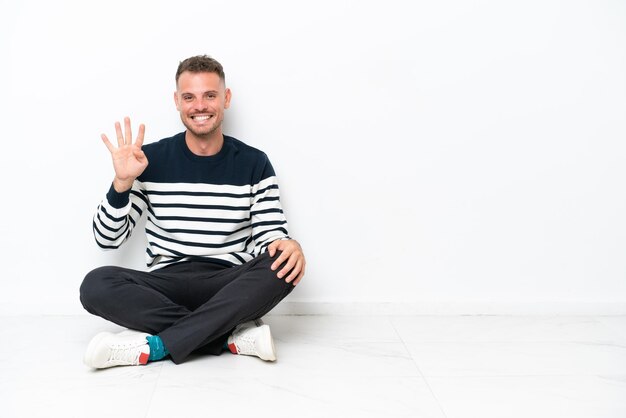 Young man sitting on the floor isolated on white background happy and counting four with fingers