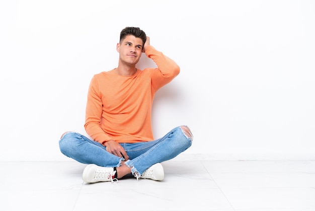 Young man sitting on the floor isolated on white background having doubts and with confuse face expression