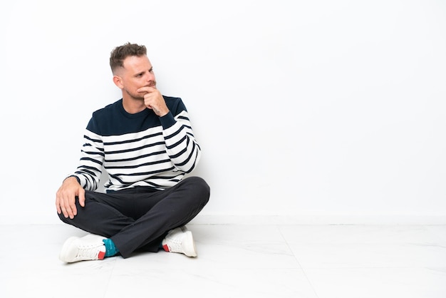 Young man sitting on the floor isolated on white background having doubts and with confuse face expression