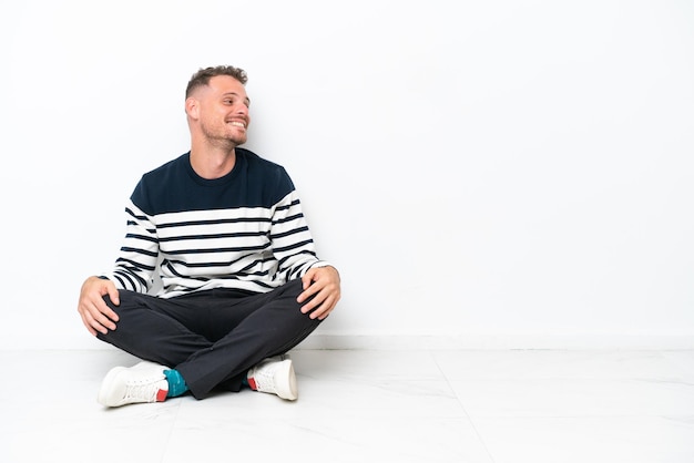 Young man sitting on the floor isolated on white background laughing in lateral position