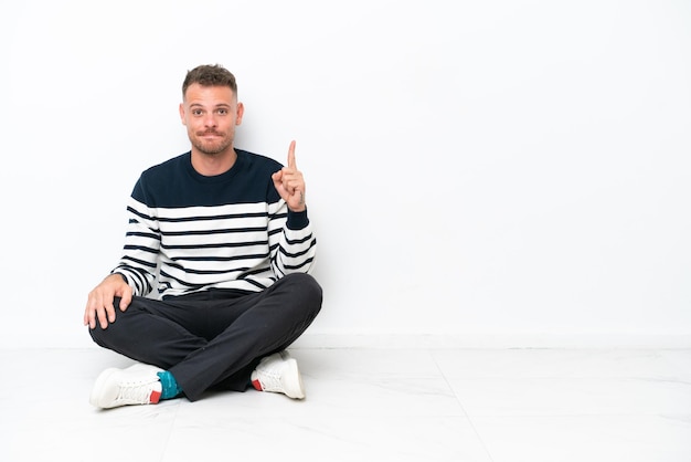 Young man sitting on the floor isolated on white background pointing with the index finger a great idea