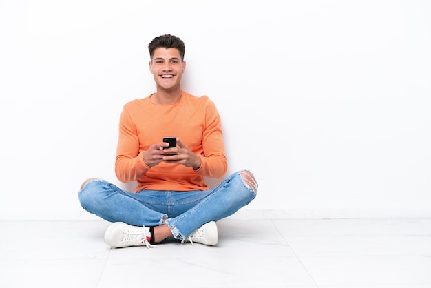 Young man sitting on the floor isolated on white background sending a message with the mobile