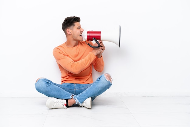 Young man sitting on the floor isolated on white background shouting through a megaphone