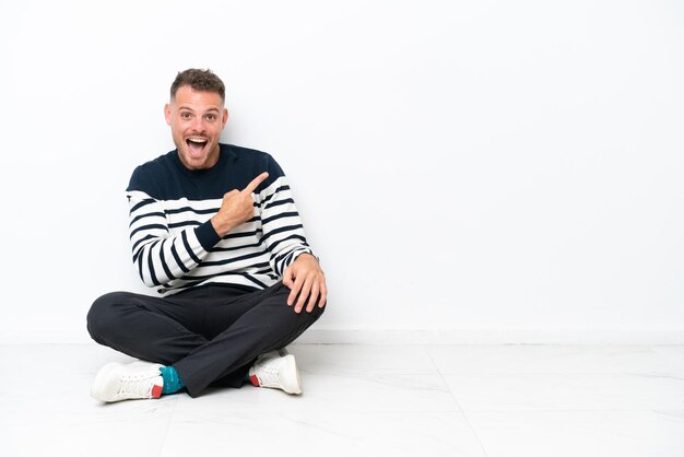 Photo young man sitting on the floor isolated on white background surprised and pointing side