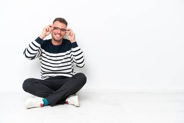 Young man sitting on the floor isolated on white background with glasses and surprised