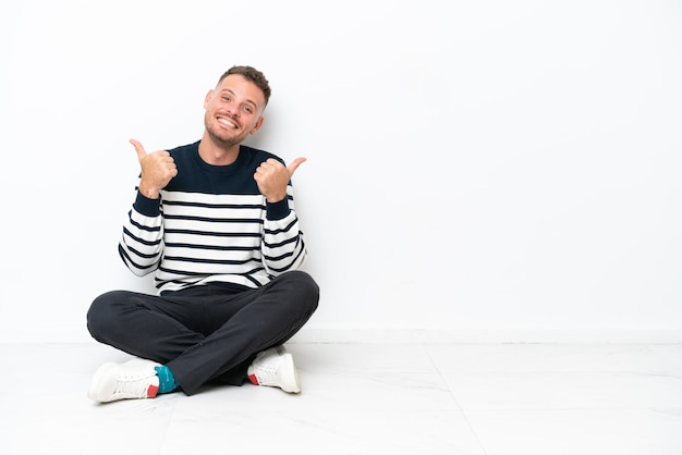 Young man sitting on the floor isolated on white background with thumbs up gesture and smiling