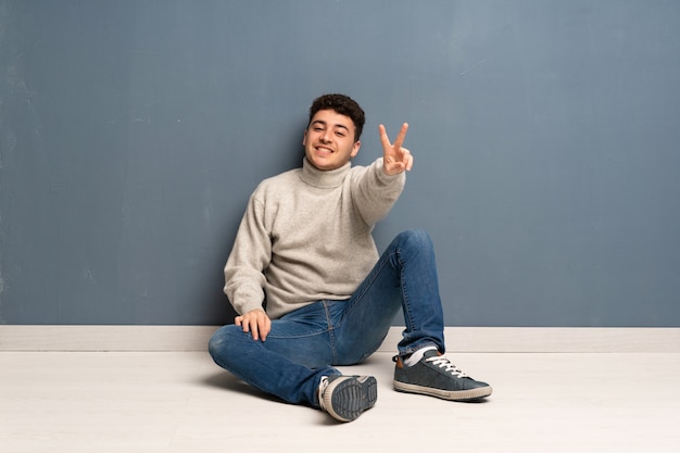 Young man sitting on the floor smiling and showing victory sign