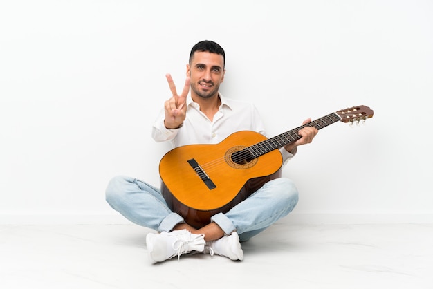 Giovane che si siede sul pavimento con la chitarra che sorride e che mostra il segno di vittoria