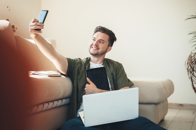 Young man sitting on the floor with a laptop and tablet making a selfie and smile