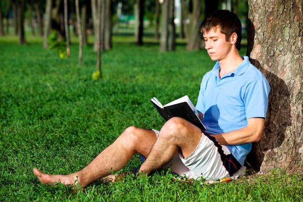 Young man sitting on green grass near tree and reading book