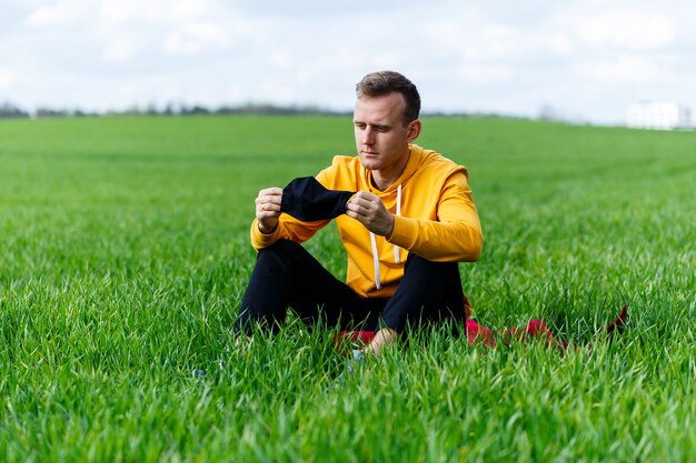 Young man sitting on green grass in a protective antiviral mask and thinking about the future. The guy is resting in the park on a sunny summer day. The concept of rest. Virus protection. Quarantine