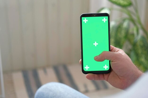 Young man sitting at home holding smartphone green mockup screen in hand Male person using chroma key mobile phone Vertical mode Touching swiping display tapping surfing Internet social media