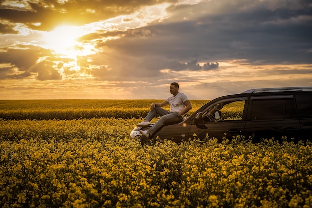 Young man sitting on the hood of a car in blooming rape field at sunset