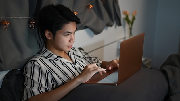 Фото Молодой человек, сидящий в спальне и использующий компьютерный ноутбук в ночное время.