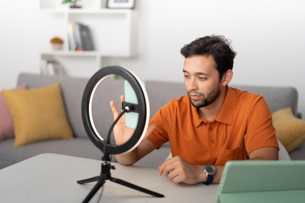 Young man sitting in a living room recording a video with his cell phone using the ring light