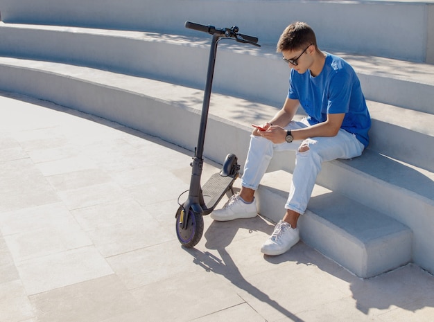 Foto giovane uomo seduto all'aperto vicino a uno scooter elettrico e usando il suo telefono