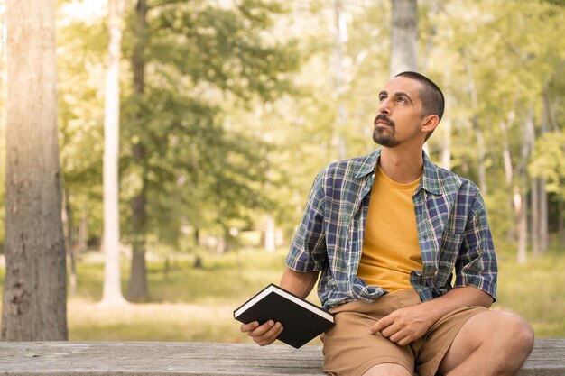 Foto giovane seduto all'aperto con un libro in mano in un parco con alberi sfondo conoscenza concetto di fede