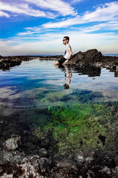 Young man sitting on rock by sea against sky