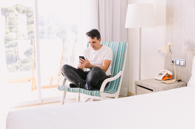 寝室のロッキングチェアに足を組んで座っている若い男が、窓から差し込む光で携帯電話を見ている休暇の接続とスマートフォンの概念