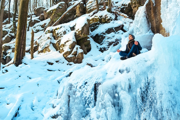 A young man sitting in the snow snowdrift in the forest and looking at the winter river