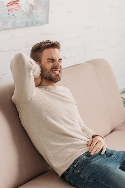 Young man sitting on sofa and suffering from pain in neck