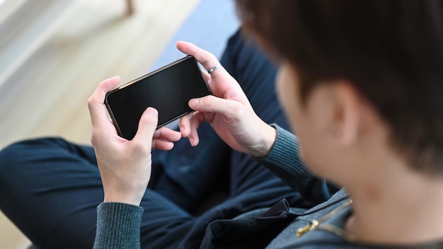 Young man sitting on a sofa with crossed legs and holding horizontal smart phone.