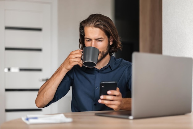 Молодой человек сидит за столом и пьет кофе, держа в руке телефон