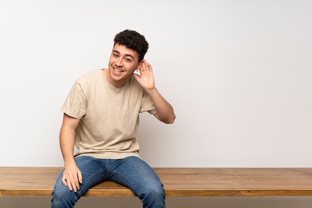 Young man sitting on table listening to something by putting hand on the ear