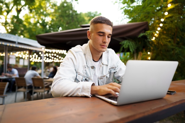 Young man sitting at table and typing on laptop keyboard while working in outdoor cafe