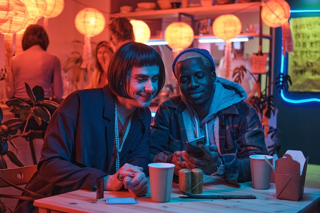 Young man sitting at the table with his boyfriend they watching something on mobile phone sitting in nightclub