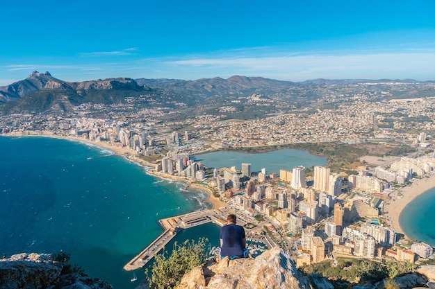 A young man sitting on top of the Penon de Ifach Natural Park in Calpe Valencia Valencian Community Spain Mediterranean sea View of the Cantal Roig and La Fossa beach