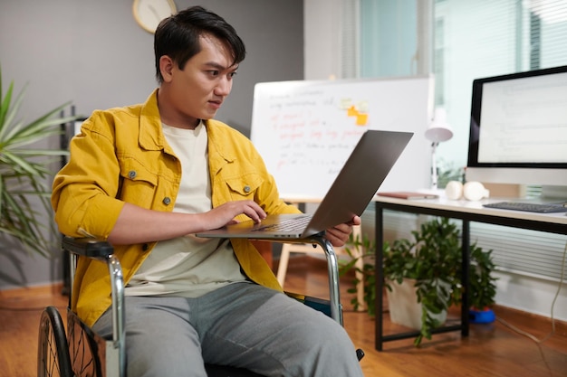 Photo young man sitting in wheelchair working on laptop in office of his own it company