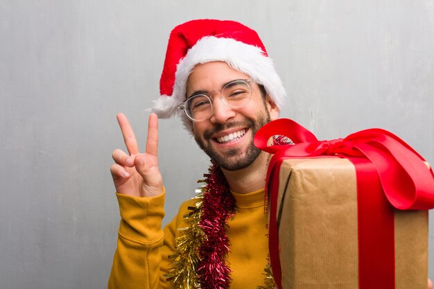 勝利のジェスチャーをしてクリスマスを祝う贈り物と一緒に座っている若い男