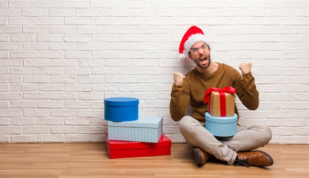 非常に怒って攻撃的な叫びクリスマスを祝うプレゼントと一緒に座っている若い男