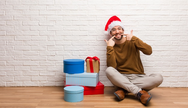 Young man sitting with gifts celebrating christmas smiles, pointing mouth